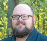 A man with glasses and a beard standing outdoors with trees in the background.