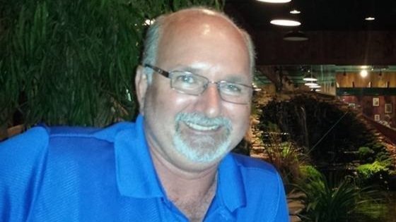 A man with glasses, a beard, and a mustache smiling, wearing a blue collared shirt, indoors with greenery and plants in the background.