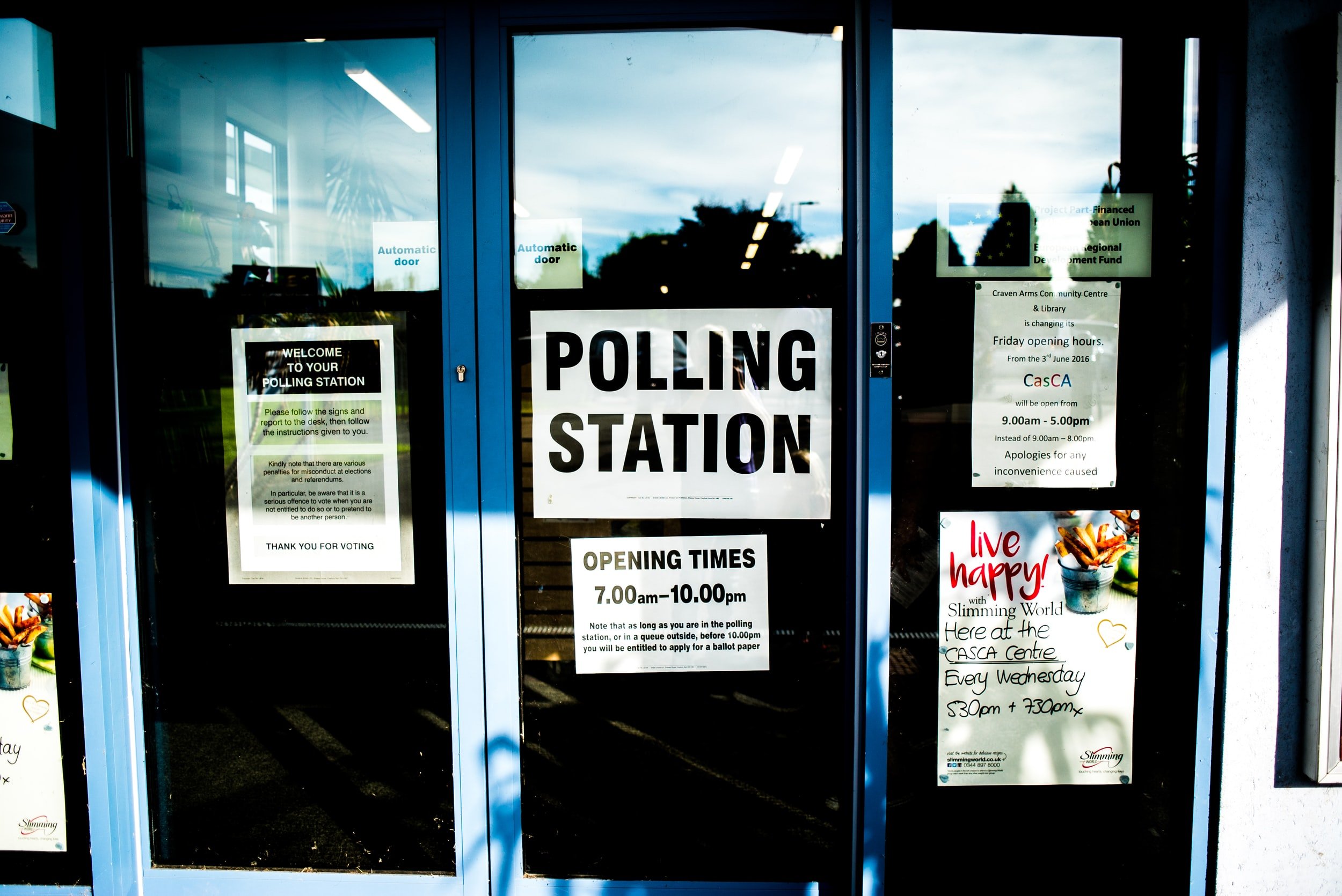Glass entrance door with signs for polling station, showing opening hours from 7:00 am to 10:00 pm, and various notices including election instructions and event promotions.