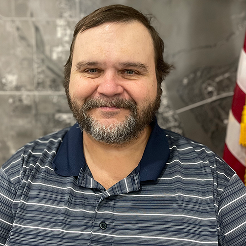 A man with dark hair and a beard smiling at the camera, wearing a striped polo shirt, standing in front of a gray wall with an American flag partially visible in the background.
