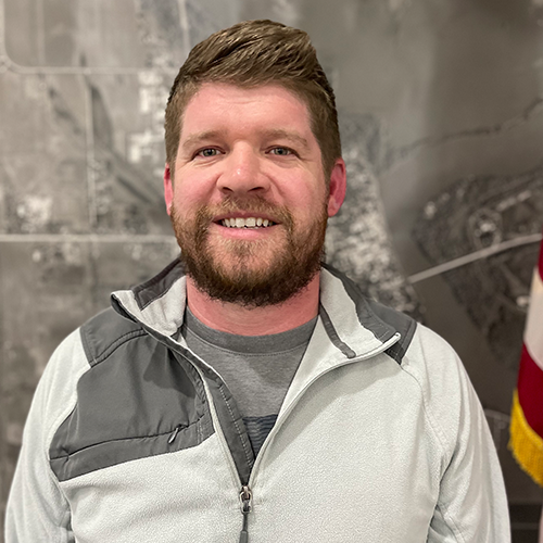 A man with a beard and short brown hair smiling, wearing a gray and black jacket, in front of a blurred map background and a partial American flag.